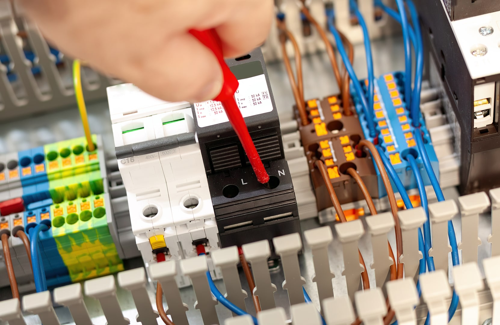 A close-up of an electrical panel with a hand using a red screwdriver on a circuit breaker. Blue and yellow wires are neatly connected, indicating precision work.