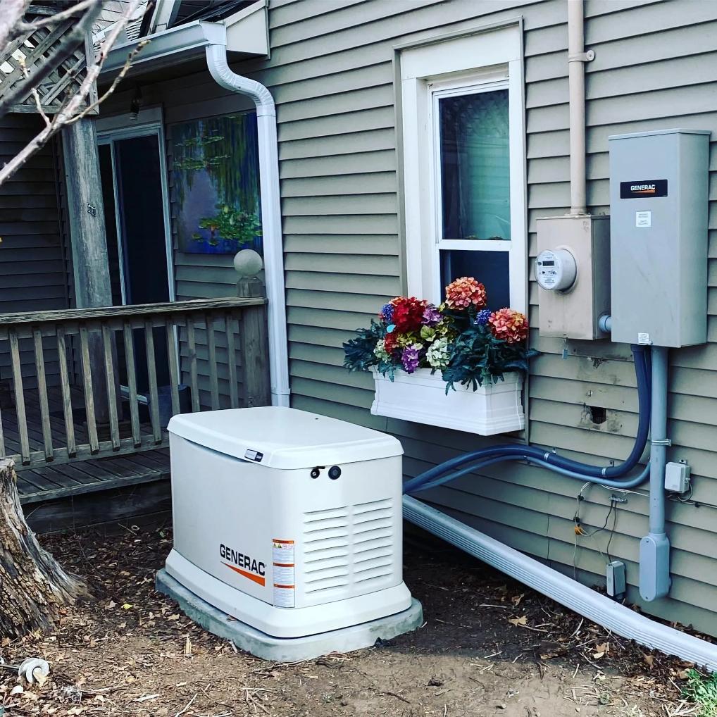 A white Generac generator is positioned outside a house with beige siding. Nearby, a window box filled with colorful flowers adds a cheerful touch. Electrical panels and wiring are visible on the wall.