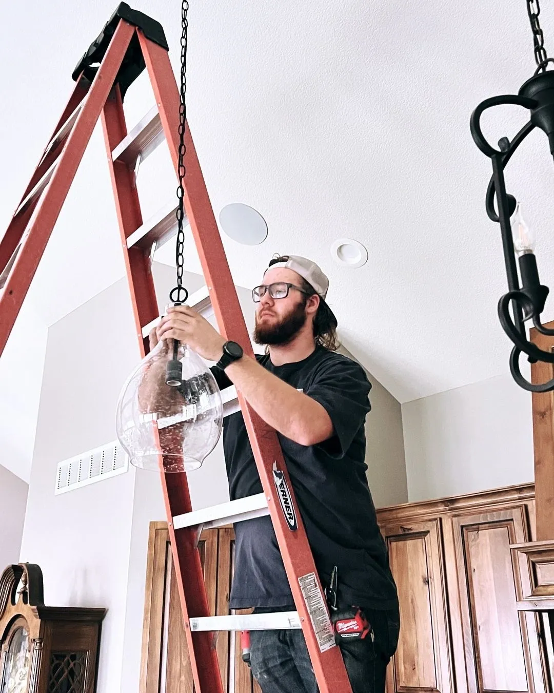 A man on a red ladder installs a glass light fixture in a room with wooden cabinets, exuding focus and precision. The setting is calm and organized.