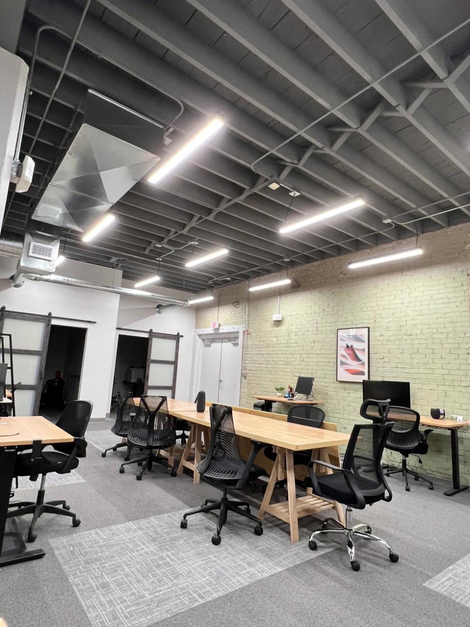 Modern office space with exposed ductwork and light fixtures on a high ceiling. Wooden desks and black chairs rest on a carpeted floor, creating a clean, airy vibe.