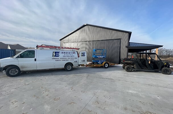 White work van with "Innovative" branding, towing a trailer with blue equipment, parked on a concrete driveway beside a large gray metal building and a black utility vehicle. Sky is clear, suggesting a calm setting.