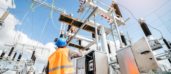 A worker in a blue helmet and orange vest adjusts equipment at an electrical substation. The sky is clear, emphasizing a focus on safety and precision.
