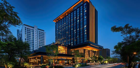 Modern hotel at dusk, featuring a tall, illuminated building with large windows, surrounded by greenery and nearby urban structures against a clear evening sky.