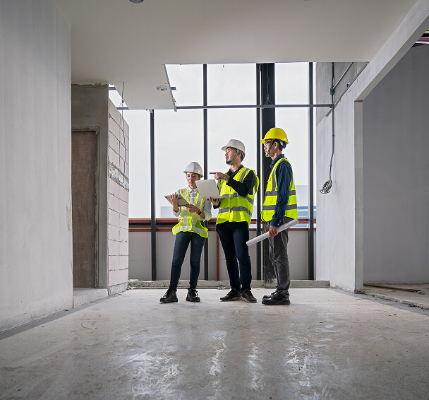 Three construction workers in hard hats and safety vests examine plans in a bright, unfinished space with large windows; the mood is focused and professional.