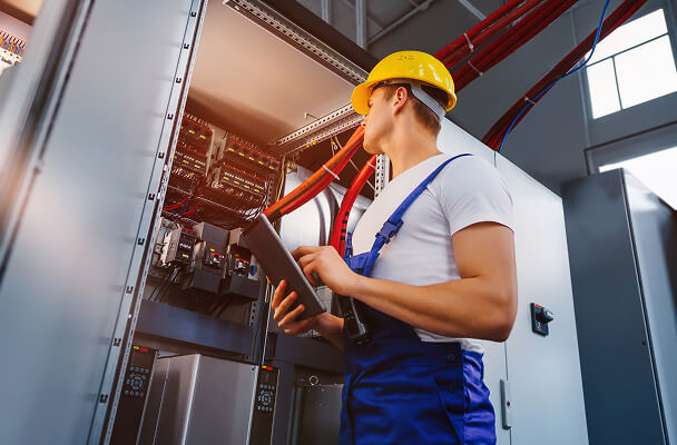 A worker in a hard hat and blue overalls examines an open electrical control panel with red cables, holding a tablet. The setting is industrial.