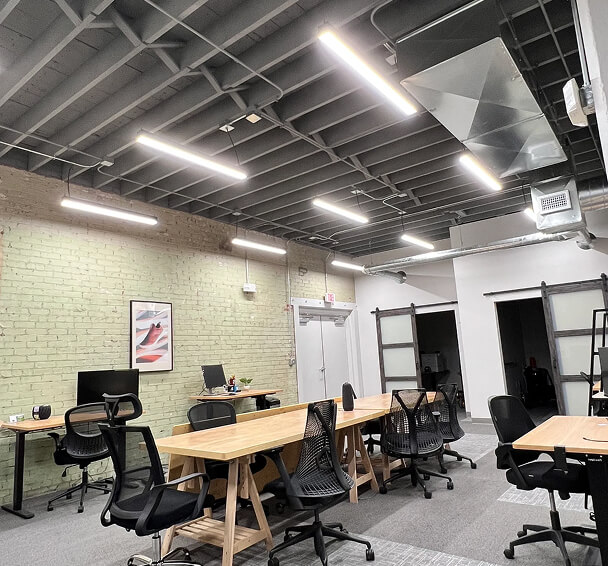 Modern office space with exposed ceiling and linear lights. It features long wooden desks, black chairs, a brick accent wall, and framed artwork.