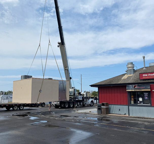 A large crane lifts a beige modular unit from a flatbed truck near a red building labeled "Old Fashioned Burger," under a partly cloudy sky. Urban setting.