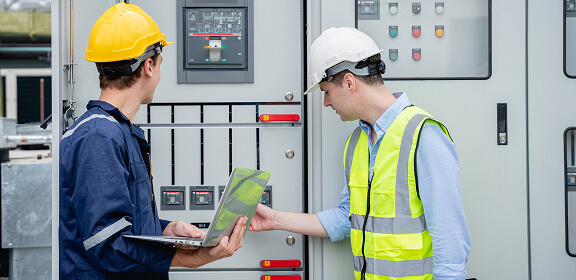 Two engineers inspecting an industrial control panel. One uses a laptop, and the other points at a switch. They wear safety gear, conveying focus.
