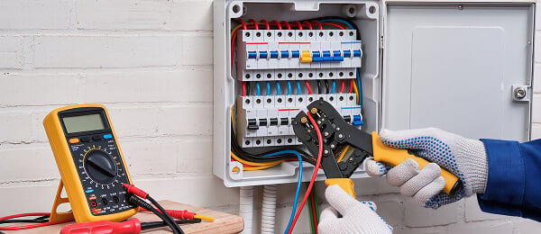 A technician in gloves uses wire strippers inside an electrical panel with colorful wires. A multimeter is on a nearby surface, monitoring voltage.