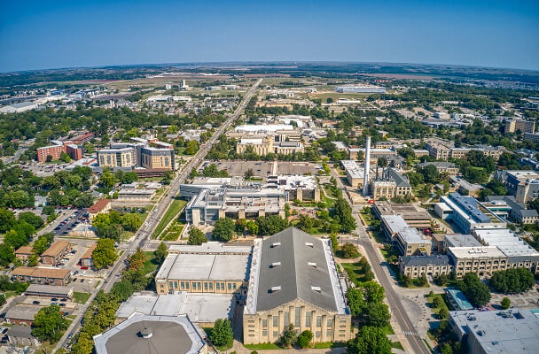 Aerial view of a cityscape featuring a mix of modern and historic buildings, tree-lined streets, and urban greenery, under a clear blue sky.