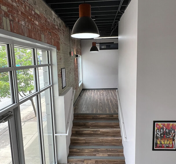 Modern hallway with exposed brick and white walls, featuring wooden floors and black pendant lights. A large window shows a tree-lined street. Urban vibe.