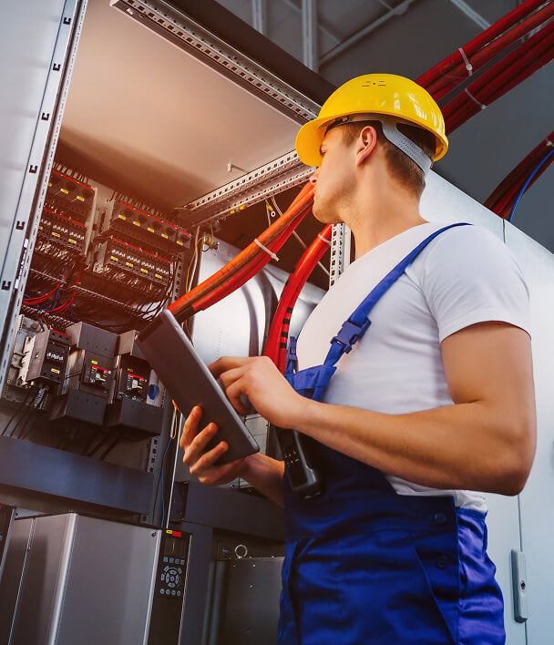 A technician in a yellow hard hat and blue overalls inspects an electrical panel with red wires, holding a clipboard, focused and attentive.